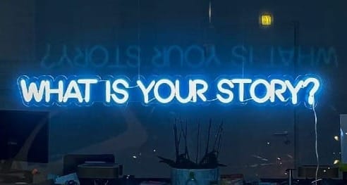 Window into an office with a neon sign that reads "What is your story?"